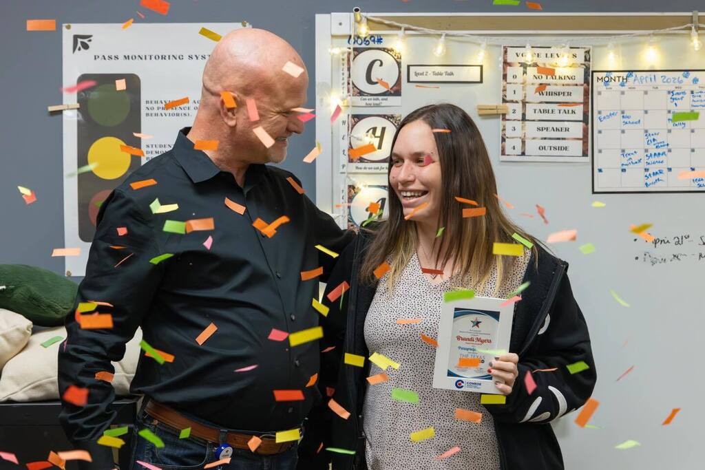 Teacher smiles holding award with Superintendent as confetti falls.