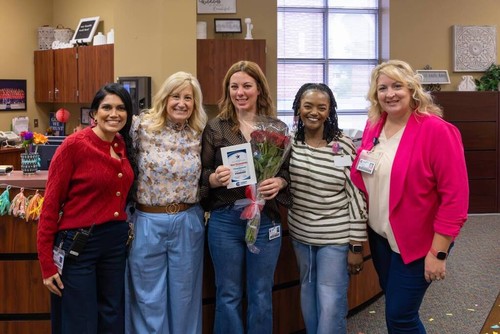 Staff member holding award plaque and flowers joined by principal and colleagues.