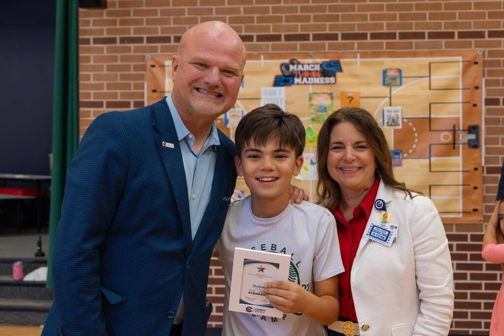 Student holding award plaque joined by Superintendent and Board President.
