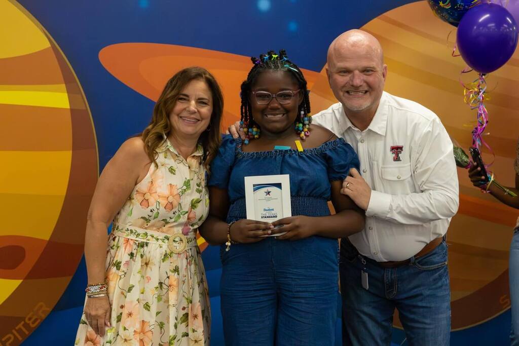 Student holding award joined by Board President and Superintendent.