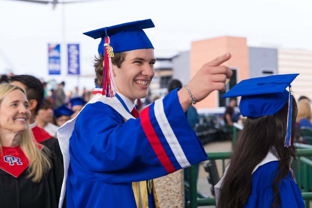 Graduate in blue robe points and smiles.