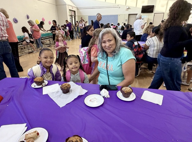 A smiling grandparent sits at a purple cover table with two young students during goodies with parents at Creighton Elementary. Muffins and pastries are on the table as families and students gather throughout the school cafeteria in the background.