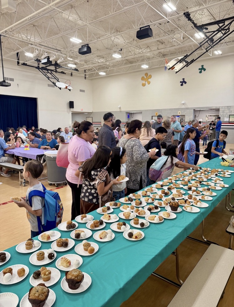 Students, grandparents, and family members gather in the Creighton Elementary cafeteria for goodies with grandparents. A long table covered in a turquoise tablecloth displays rows of plates filled with muffins, pastries, and other breakfast treats.