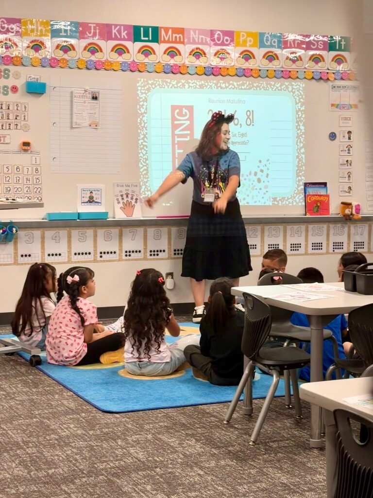 A teacher leads a morning meeting while elementary students sit together on a classroom rug. A projected slide behind the teacher displays a greeting activity, while colorful alphabet and rainbow decorations line the classroom wall.