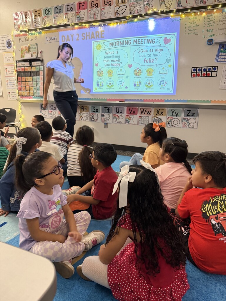 A teacher stands in front of a group of elementary students seated on the classroom floor during a morning meeting. The projected bilingual activity asks students, “What is something that makes you happy?” as a student raises her hand to participate.