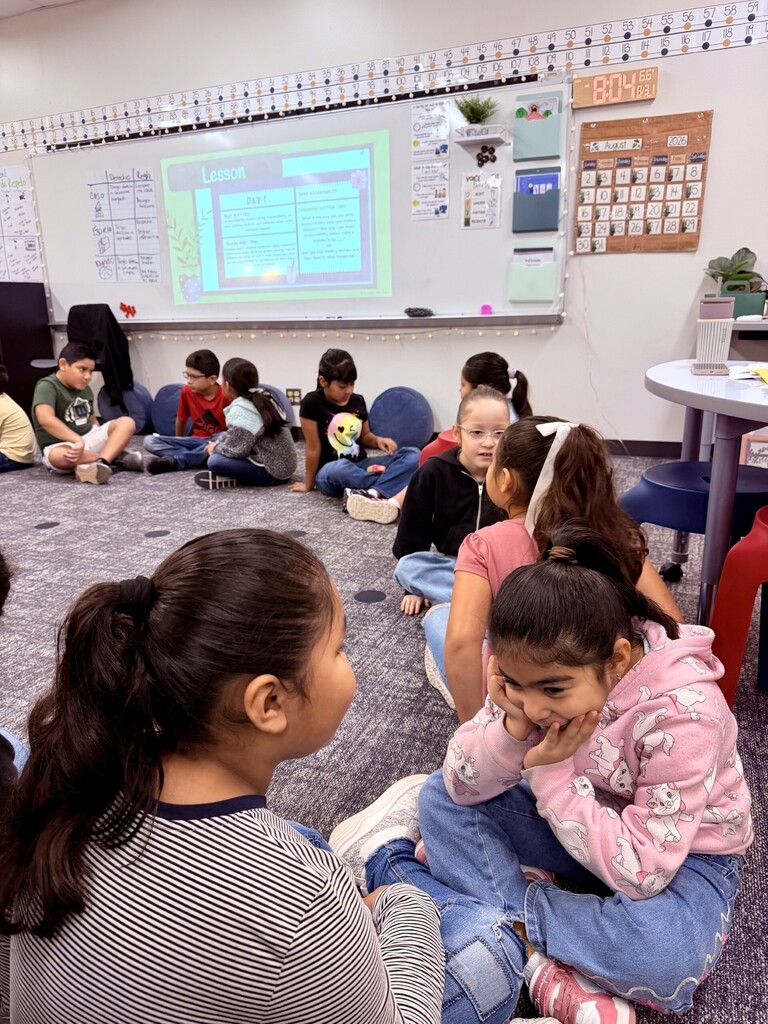 Elementary students sit in pairs on the classroom floor and face one another during a partner discussion activity. Several pairs are talking and listening to each other while a lesson slide is projected at the front of the classroom.
