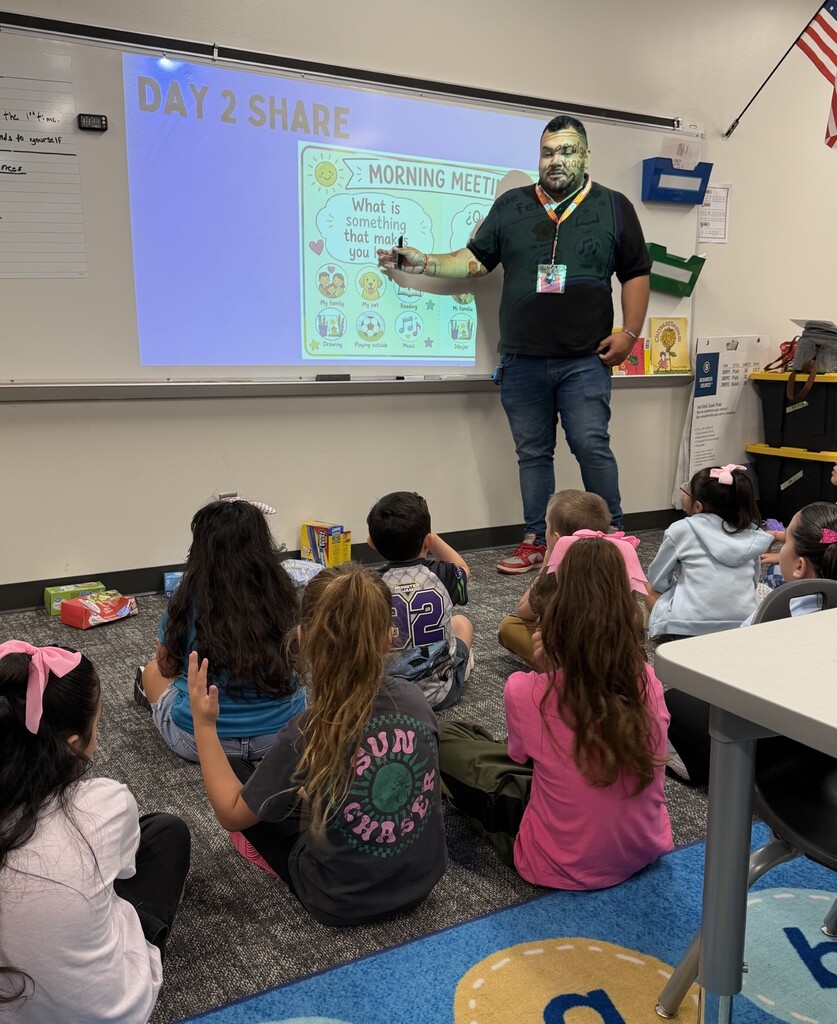 A teacher stands in front of a group of elementary students seated on the classroom floor during a morning meeting. The projected bilingual activity asks students, “What is something that makes you happy?” as a student raises her hand to participate.