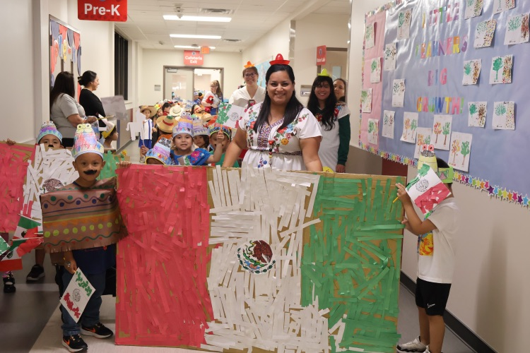 Pre-K students and teachers celebrate Mexican heritage with colorful outfits, handmade decorations, and a large Mexican flag during a school parade.