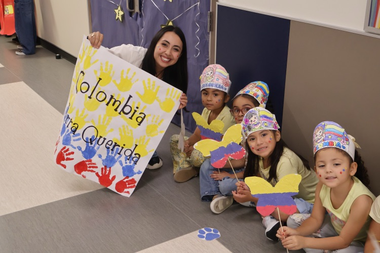 A teacher and four students celebrate Colombian heritage with colorful handmade hats, butterfly decorations, and a Colombia-themed poster.