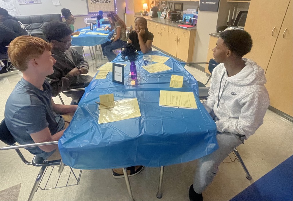 Students sitting or standing around a table discussing various topics.