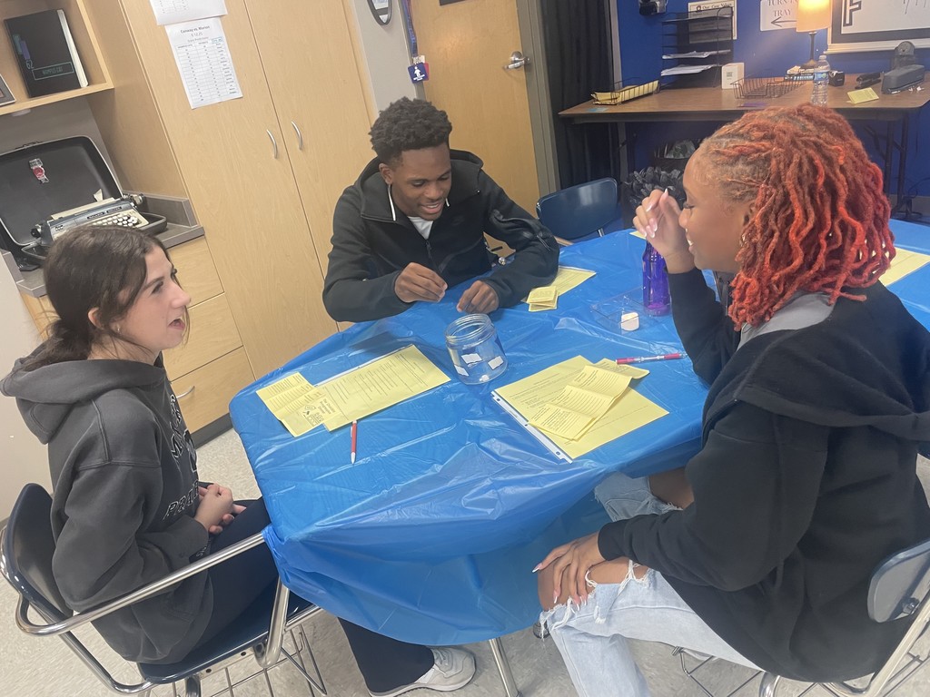 Students sitting or standing around a table discussing various topics.