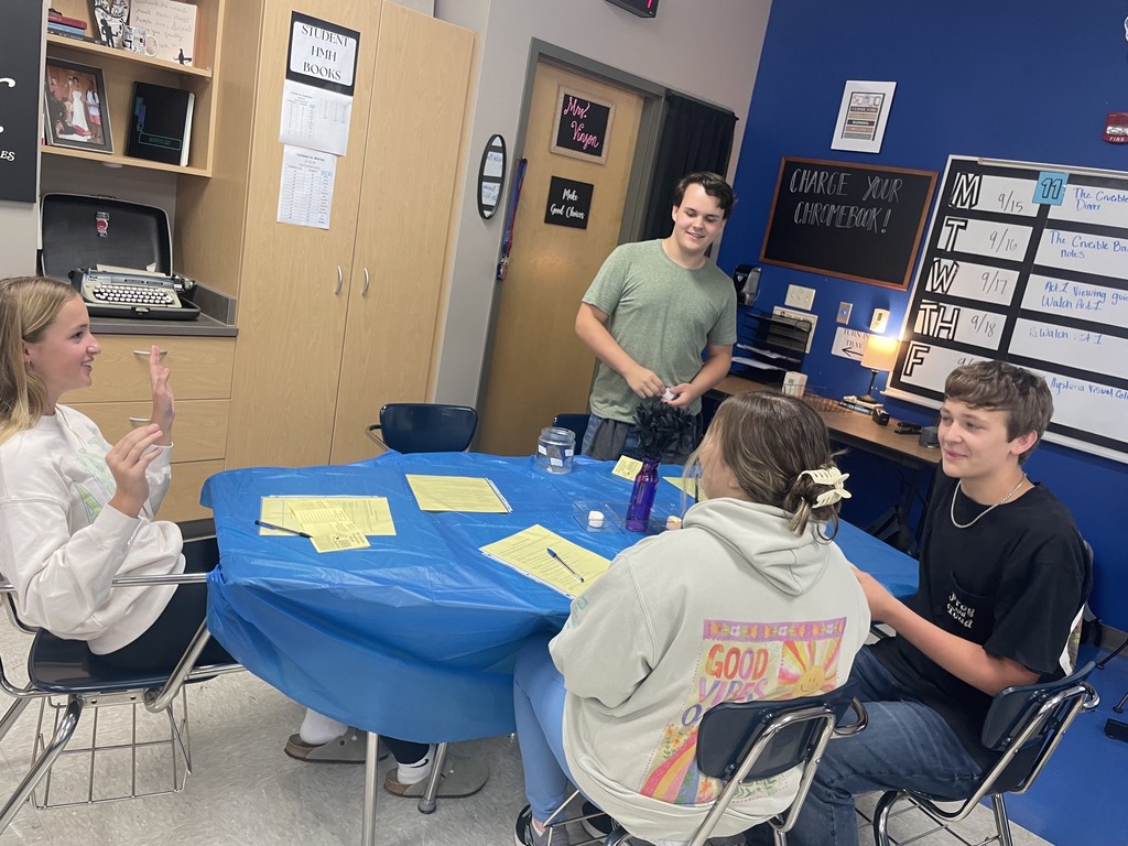 Students sitting or standing around a table discussing various topics.