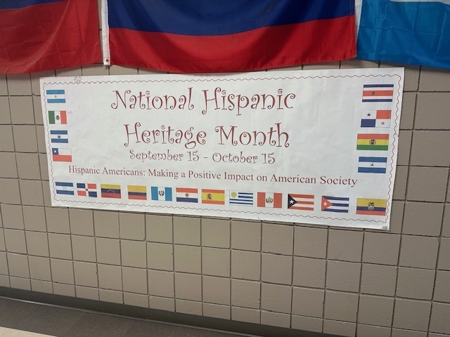 National Hispanic Heritage Month banner hung on a brick wall surrounded by a border of various flags from different countries.