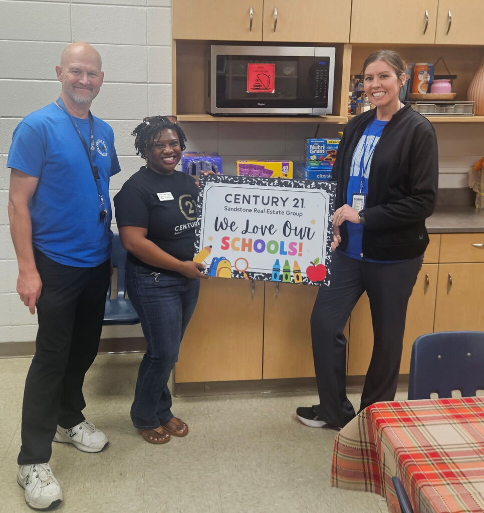 Three adults standing in a staff breakroom holding a framed sign that reads "CENTURY 21 Sandstone Real Estate Group - We Love Our SCHOOLS!" surrounded by donated boxes of snacks like Nutri-Grain bars.