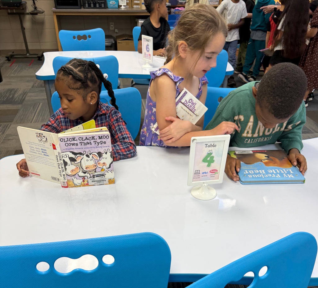 Three young elementary students sitting at a white classroom table labeled "Table 4." On the left, a girl in a plaid shirt reads the book Click, Clack, Moo: Cows That Type. In the middle, a girl in a purple floral dress stands/leans over with a book under her arm. On the right, a boy in a green sweatshirt leans down to read My Precious Little Bear.