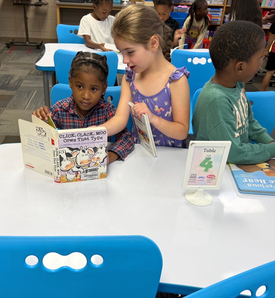 Three young students gathered around a white classroom table labeled "Table 4." A young girl in a purple floral dress leans in to look at the book Click, Clack, Moo: Cows That Type held by a classmate wearing a plaid shirt. A boy in a green sweatshirt sits beside them looking away.