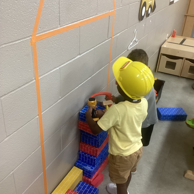 two children wearing safety gear constructing a house using cardboard blocks and tools