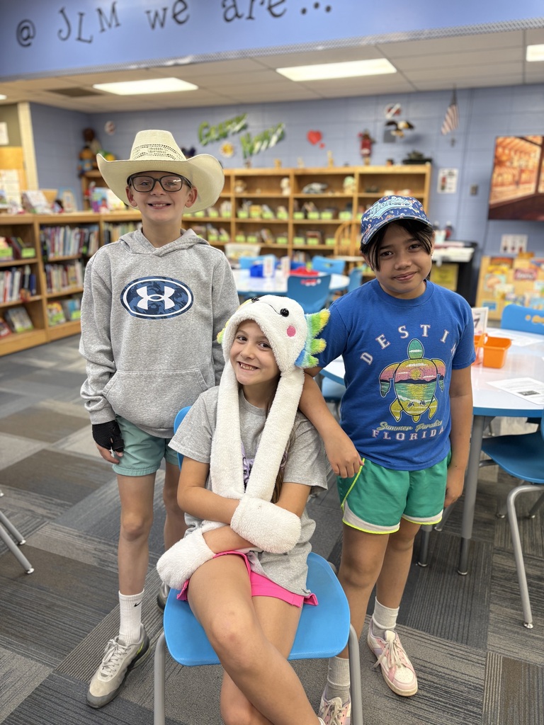 Three kids in a library. A boy and a girl wearing hats and a girl wearing a dinosaur hat.