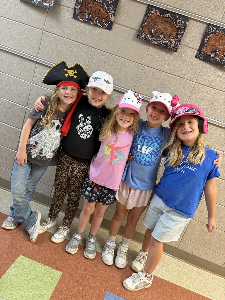 Five young girls wearing hats, standing in a hallway, smiling, with one hugging another.