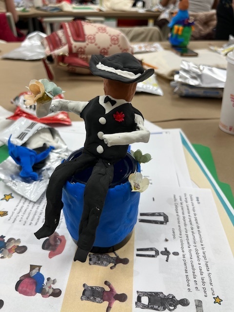 Clay sculpture of a person in a black outfit with a hat, sitting on a blue object, holds a flower. Background shows art supplies and papers on a table.