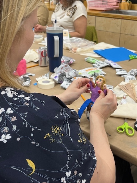 A woman in a floral shirt crafts a small doll at a cluttered table with art supplies, conveying creativity and focus in a lively, workshop setting.