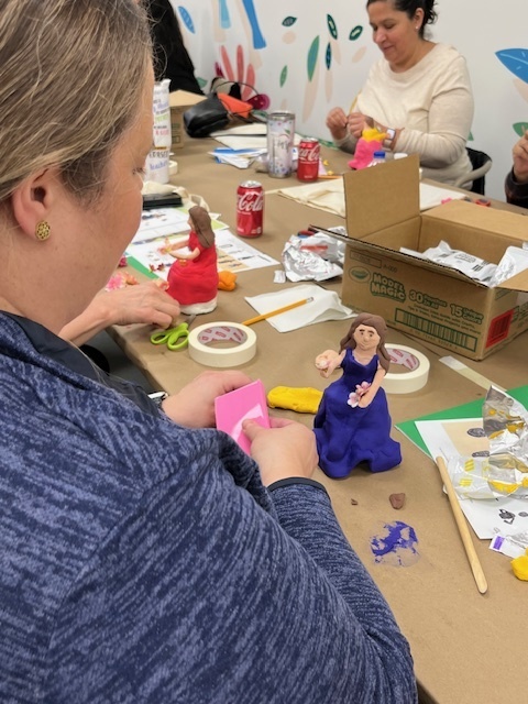 A group of people craft small, colorful figures from clay at a table. The atmosphere is creative and focused, with art supplies scattered around.