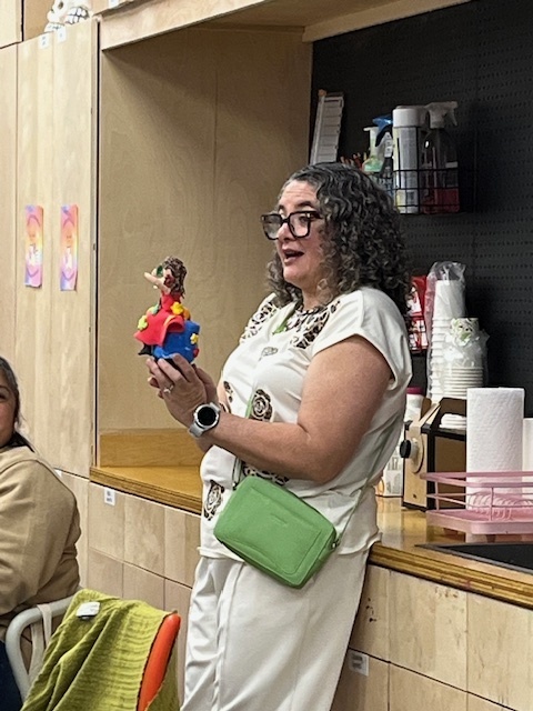 Woman holding a colorful figurine, wearing glasses, a cream outfit, and a green bag. She's speaking animatedly in a cozy room, with supplies on shelves.