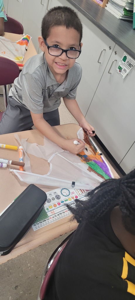 A young boy with glasses smiles while decorating a kite with colorful markers at a classroom table. The setting is creative and joyful.