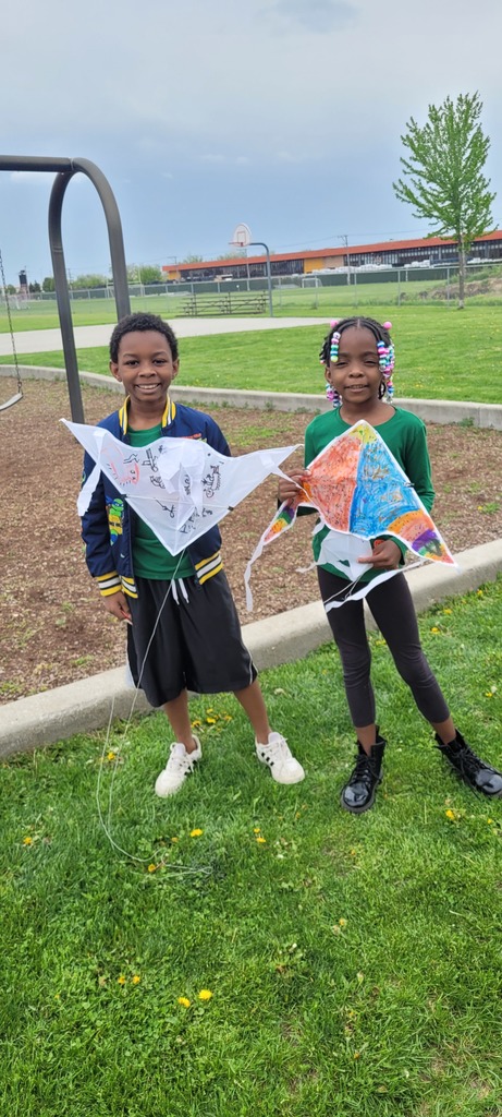 Two smiling children in a park hold colorful kites, standing on green grass. A playground structure and trees are visible in the background.
