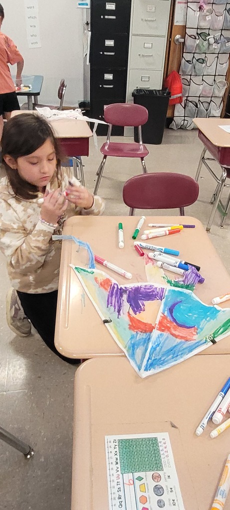 A young girl sits at a classroom desk coloring a paper kite with bright markers. The desk is scattered with art supplies, creating a creative and playful atmosphere.