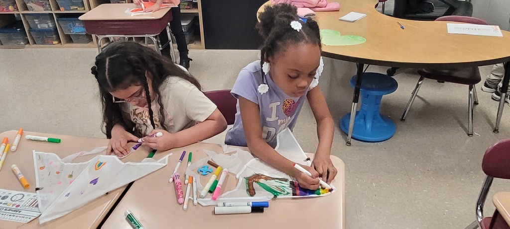 Two young girls sit at a classroom table, focused on coloring cloth bags with markers. The setting is creative and engaging.