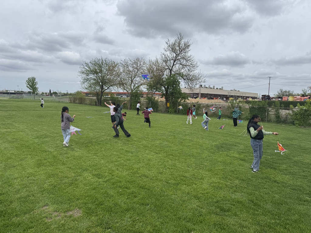 Children fly colorful kites on a lush green field under a cloudy sky, surrounded by trees and a distant industrial building, conveying joy and freedom.