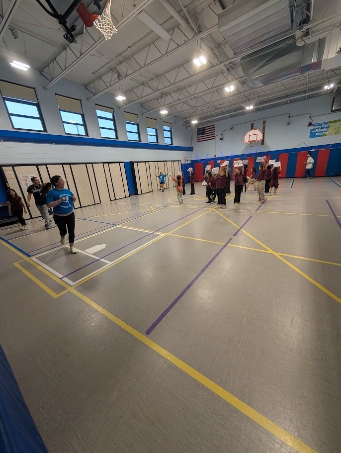 Children in maroon shirts hold boxes during an indoor school activity in a gym. A basketball hoop and an American flag are visible in the background.