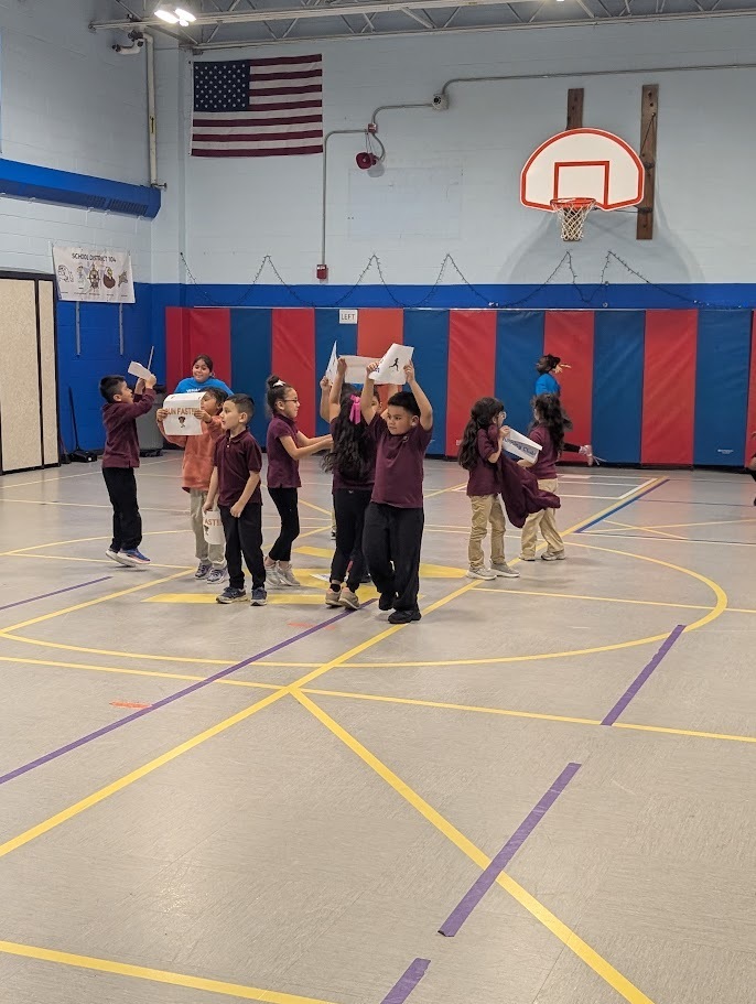 Children in maroon shirts hold boxes during an indoor school activity in a gym. A basketball hoop and an American flag are visible in the background.