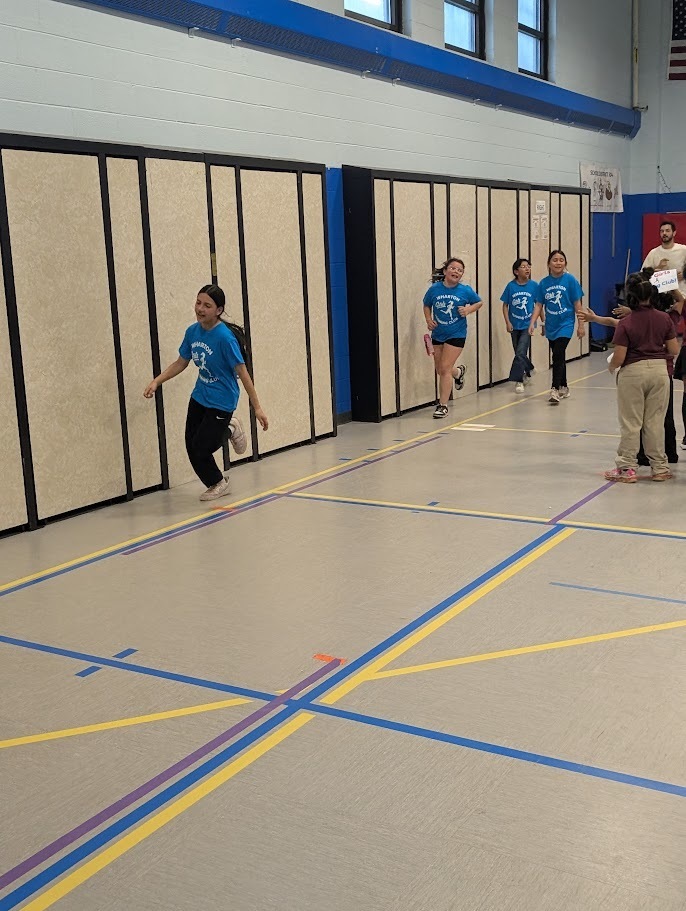 A group of girls in blue shirts runs joyfully indoors near gym walls, with colorful taped lines on the floor. The mood is energetic and lively.
