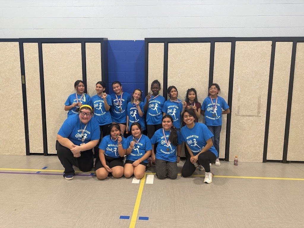 A group of children and two adults pose in a gym, smiling and wearing blue "Wharton Running Club" shirts. Some hold medals, conveying a sense of achievement and joy.