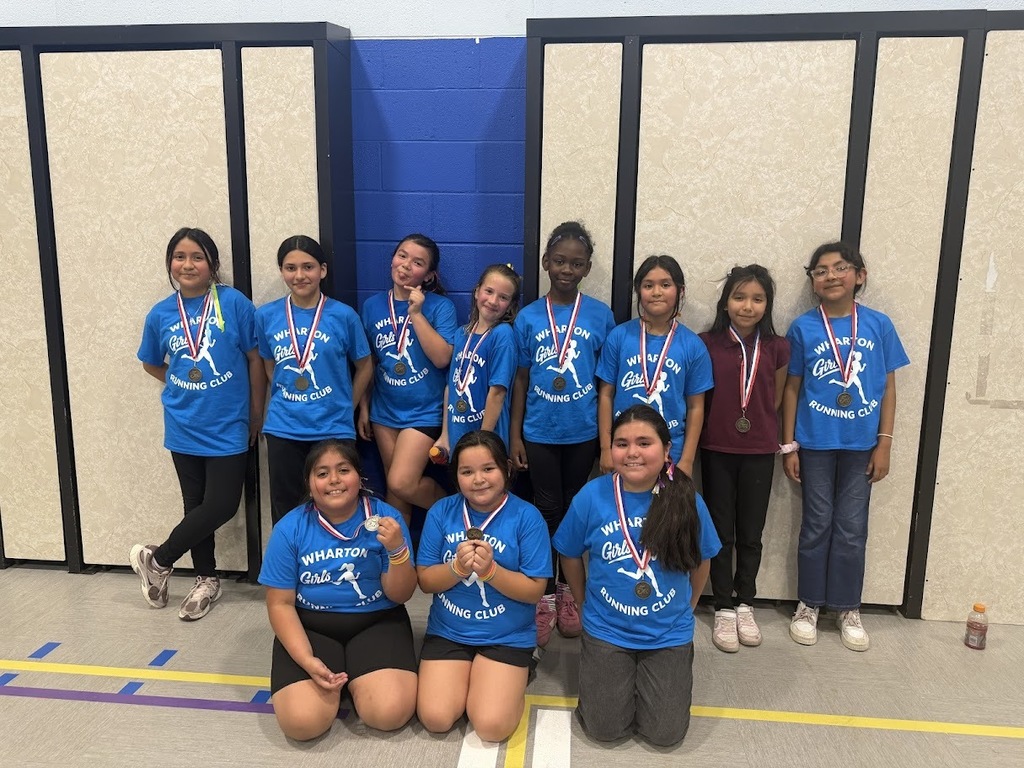 A group of young girls in blue "Wharton Girls Running Club" shirts pose indoors, proudly displaying their medals. They exude joy and teamwork.