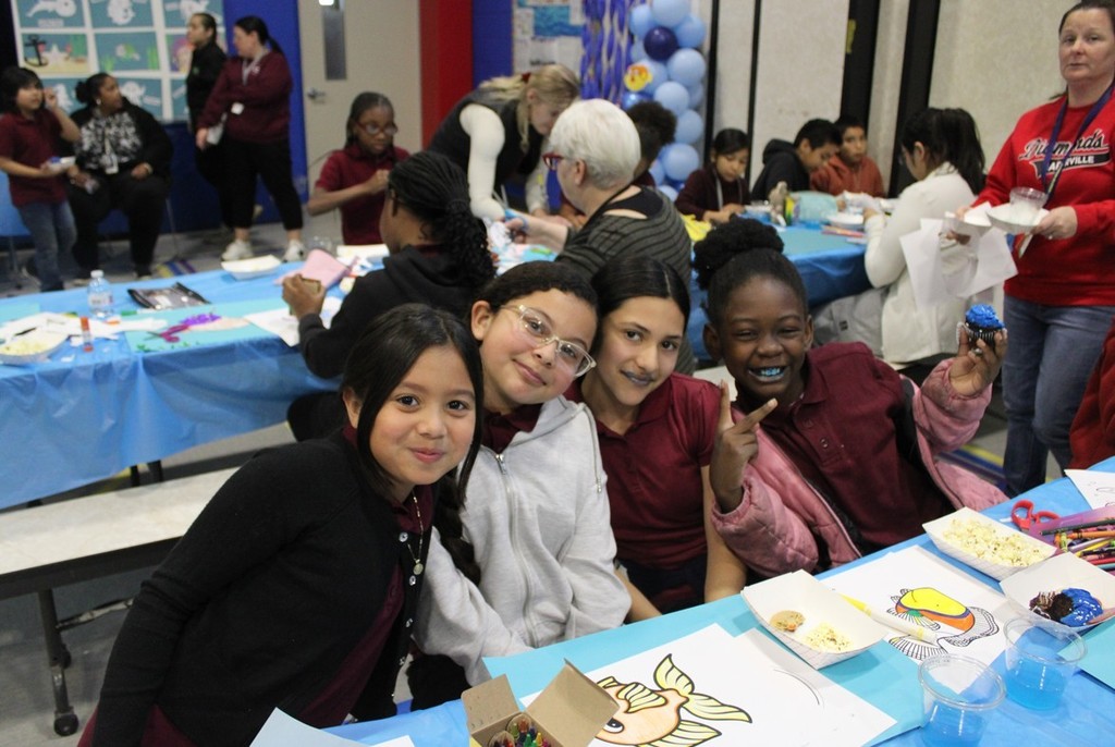 A group of four smiling children in a brightly decorated room with craft materials and snacks on a blue-covered table, conveying a joyful atmosphere.