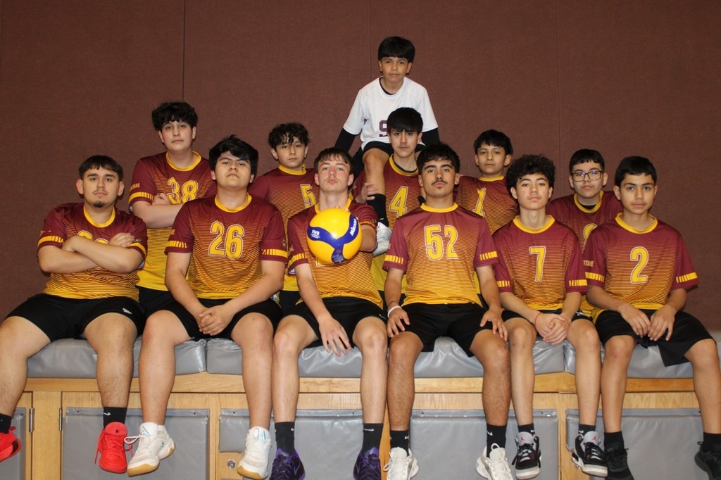 A youth volleyball team poses indoors, seated on a bench. They wear maroon and gold uniforms, with one player holding a volleyball, exuding confidence and camaraderie.