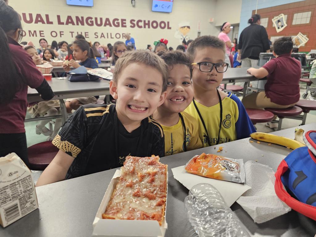 Three children smile at a cafeteria table with pizza, a snack, and a banana. Other kids are in the background at "Dual Language School." The atmosphere is cheerful and lively.