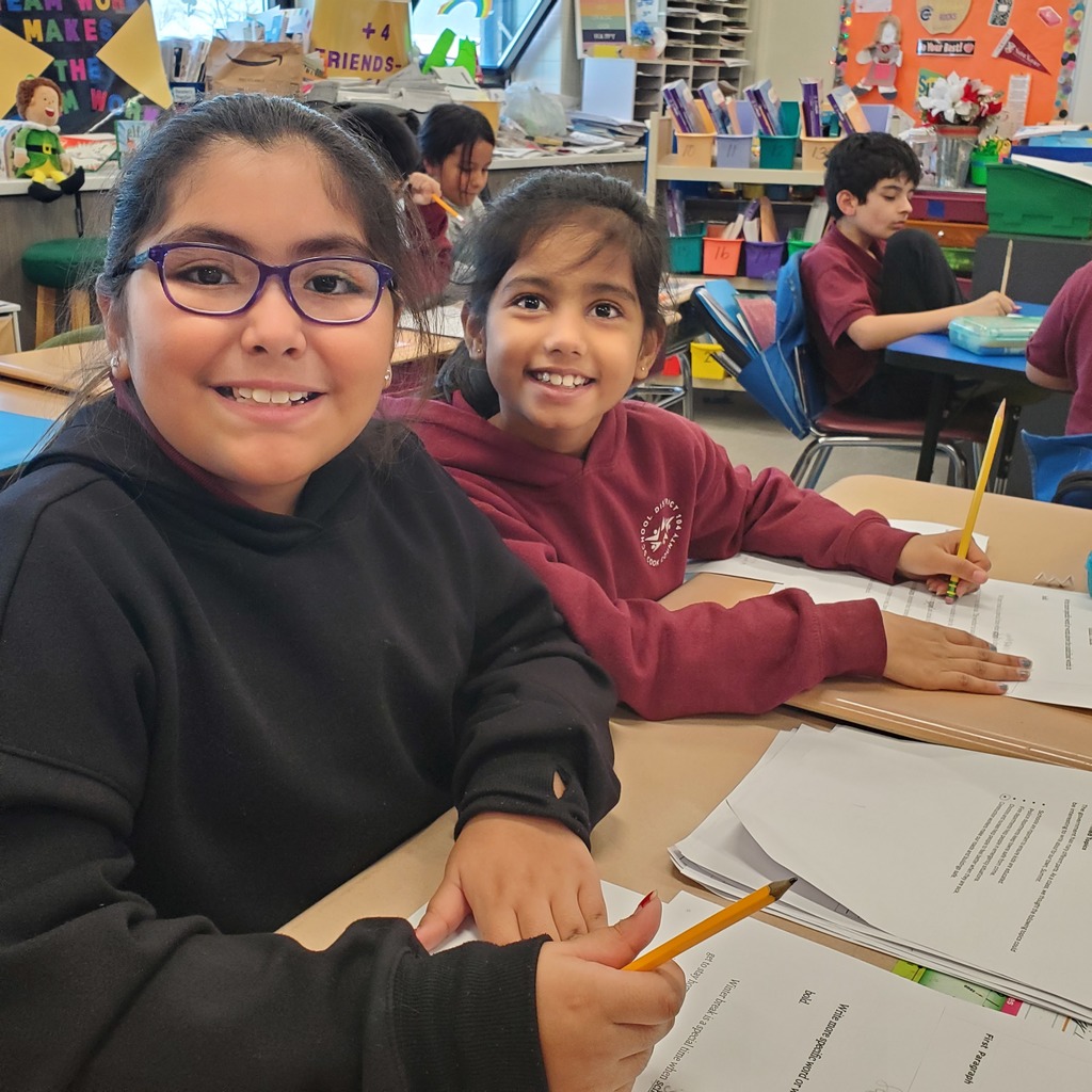 Two smiling girls at a classroom table, holding pencils and working on papers. Shelves with colorful books and posters are visible in the background.