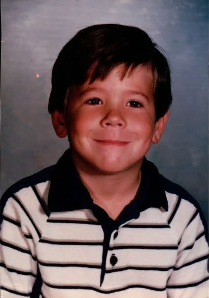 A young boy smiles warmly, wearing a striped polo shirt. His hair is neatly combed, and the background is a soft blue, giving a cheerful, nostalgic feel.