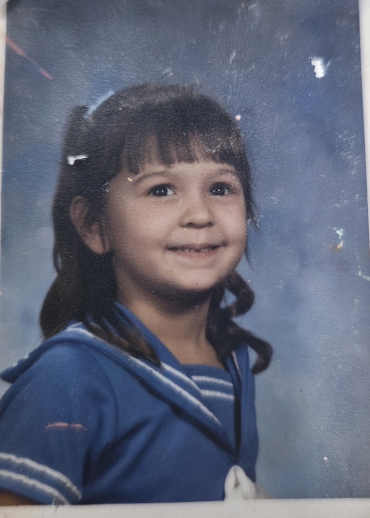 Young child with brown hair smiles softly, wearing a blue sailor-style dress against a mottled blue background, conveying a sense of innocence.