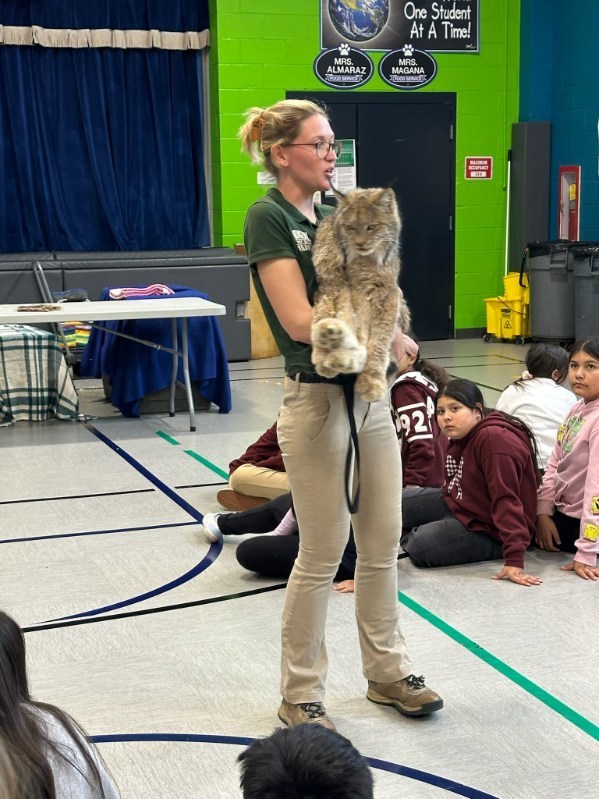 A woman holding a lynx stands in a gymnasium, speaking to seated children. The atmosphere is educational and engaging, with kids focused intently.