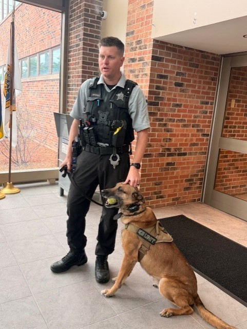 A police officer stands indoors next to a sitting police dog wearing a vest labeled "K-9 Unit." The dog holds a ball in its mouth. Brick wall background.