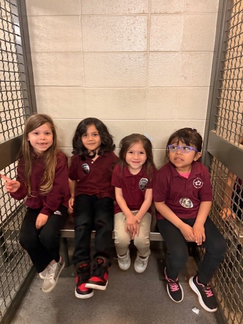 Four young children sit closely on a bench, all wearing matching maroon shirts. They smile softly, projecting a sense of camaraderie and warmth.