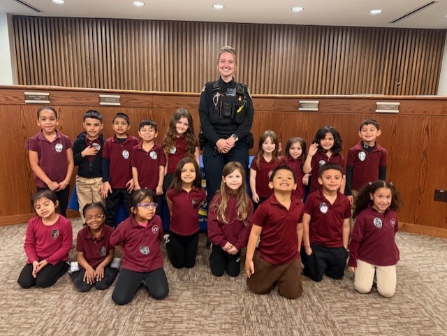 A smiling police officer stands with a group of schoolchildren in maroon uniforms. The children, some kneeling, others standing, are posing happily indoors.