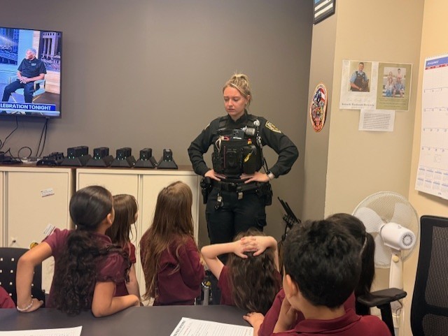 A police officer in uniform speaks to attentive young children seated around a table in a classroom. The room is equipped with a TV and electronic devices.