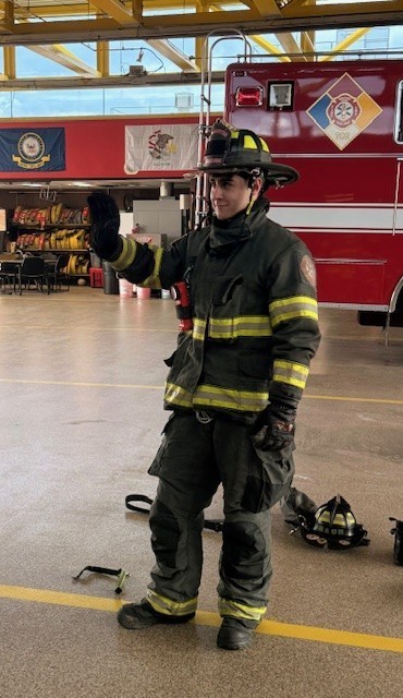 A firefighter in full gear stands inside a fire station, gesturing with one hand. In the background, a red fire truck and flags are visible.