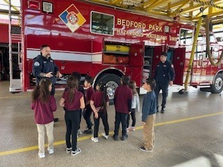 Firefighters in uniform stand by a red Bedford Park hazardous materials truck, talking to a group of elementary school children wearing red shirts, inside a fire station.